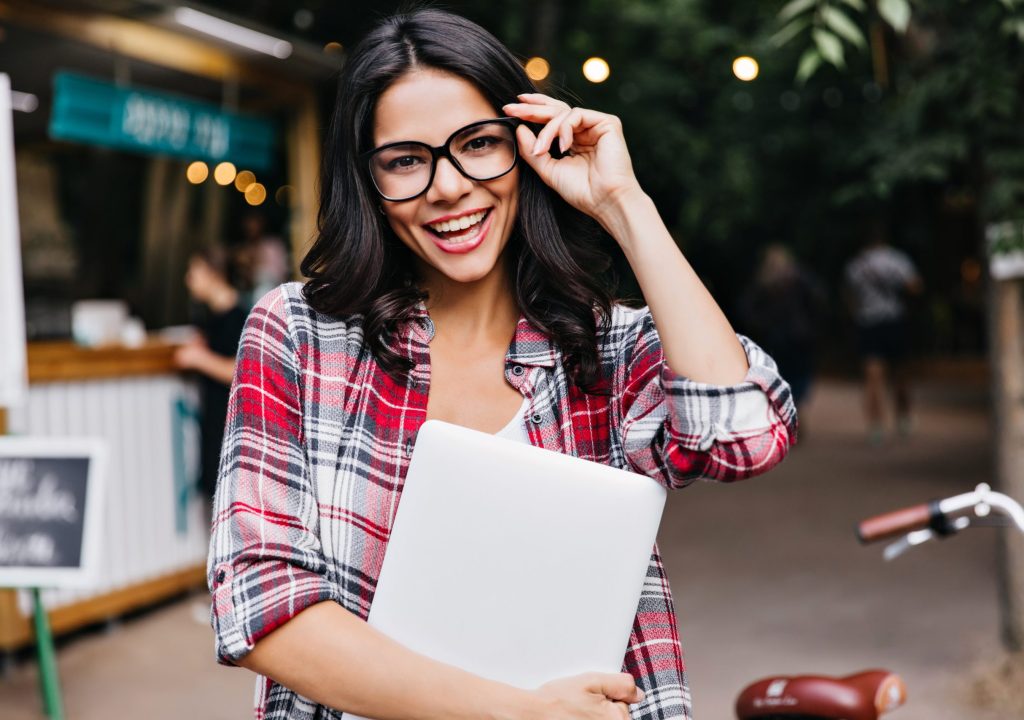 Blissful dark-haired girl with laptop touching her glasses. Outdoor portrait of happy latin female freelancer.